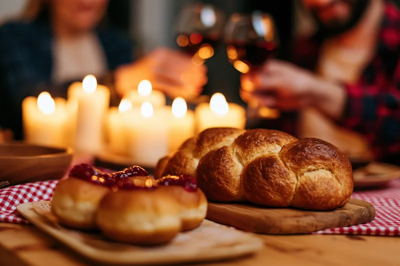 Braided challah and jam-covered pastries on a wooden board at a candlelit dinner table with a couple toasting in the background