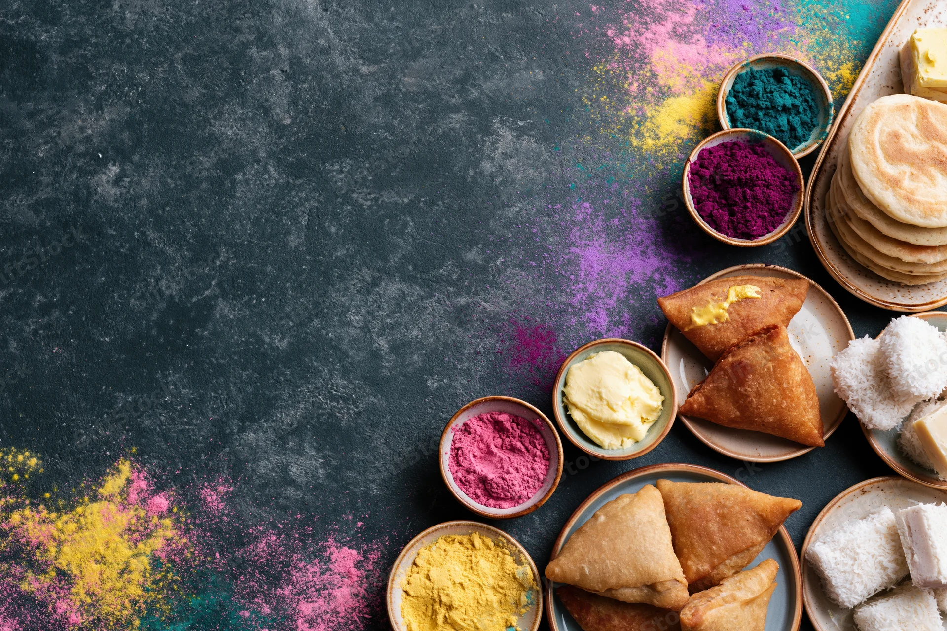 Holi festival food spread arranged along the right edge of a dark surface with colorful gulal powder and traditional sweets