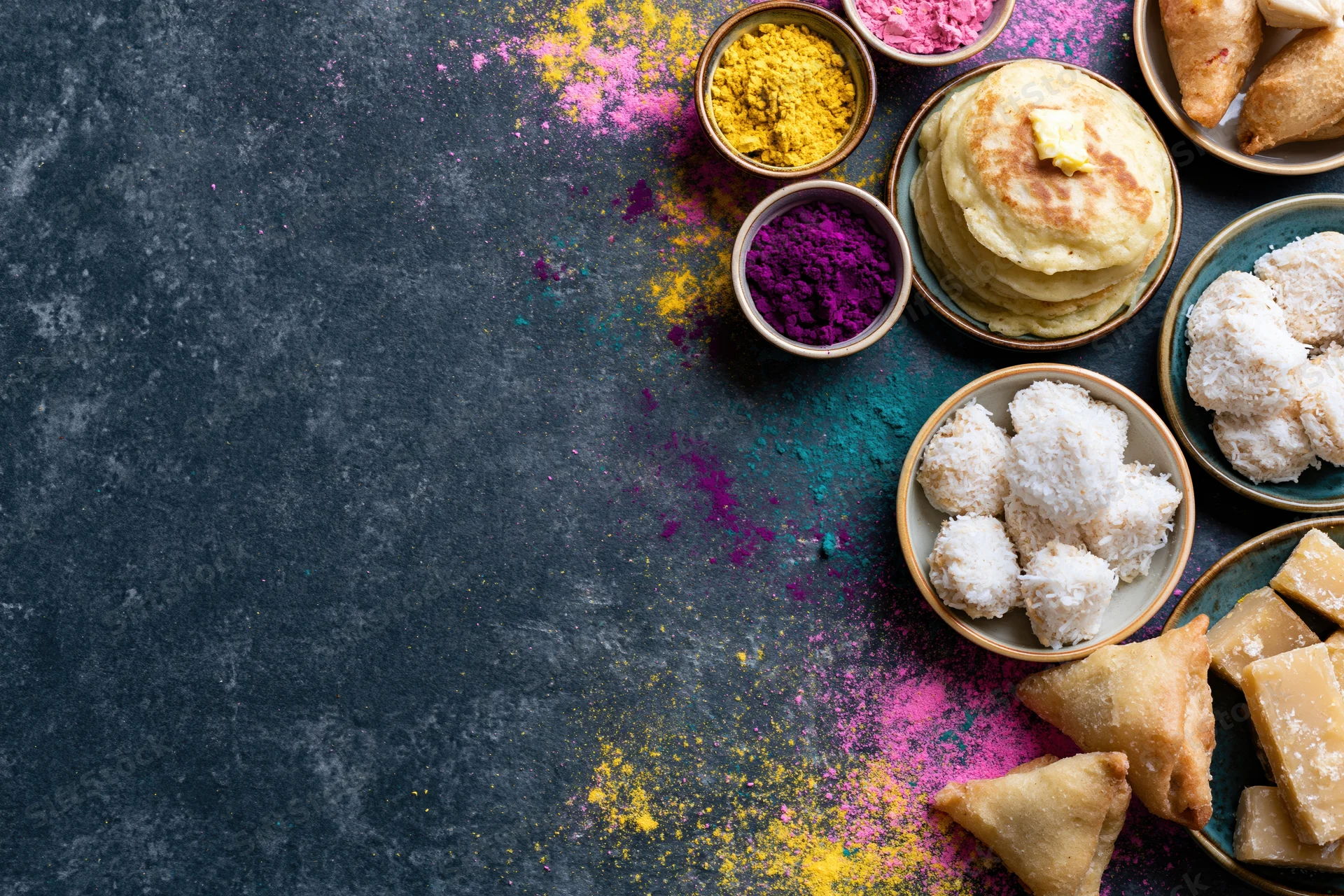 Holi festival food spread arranged along the right edge of a dark textured table with colorful gulal powder and traditional sweets in bowls and plates