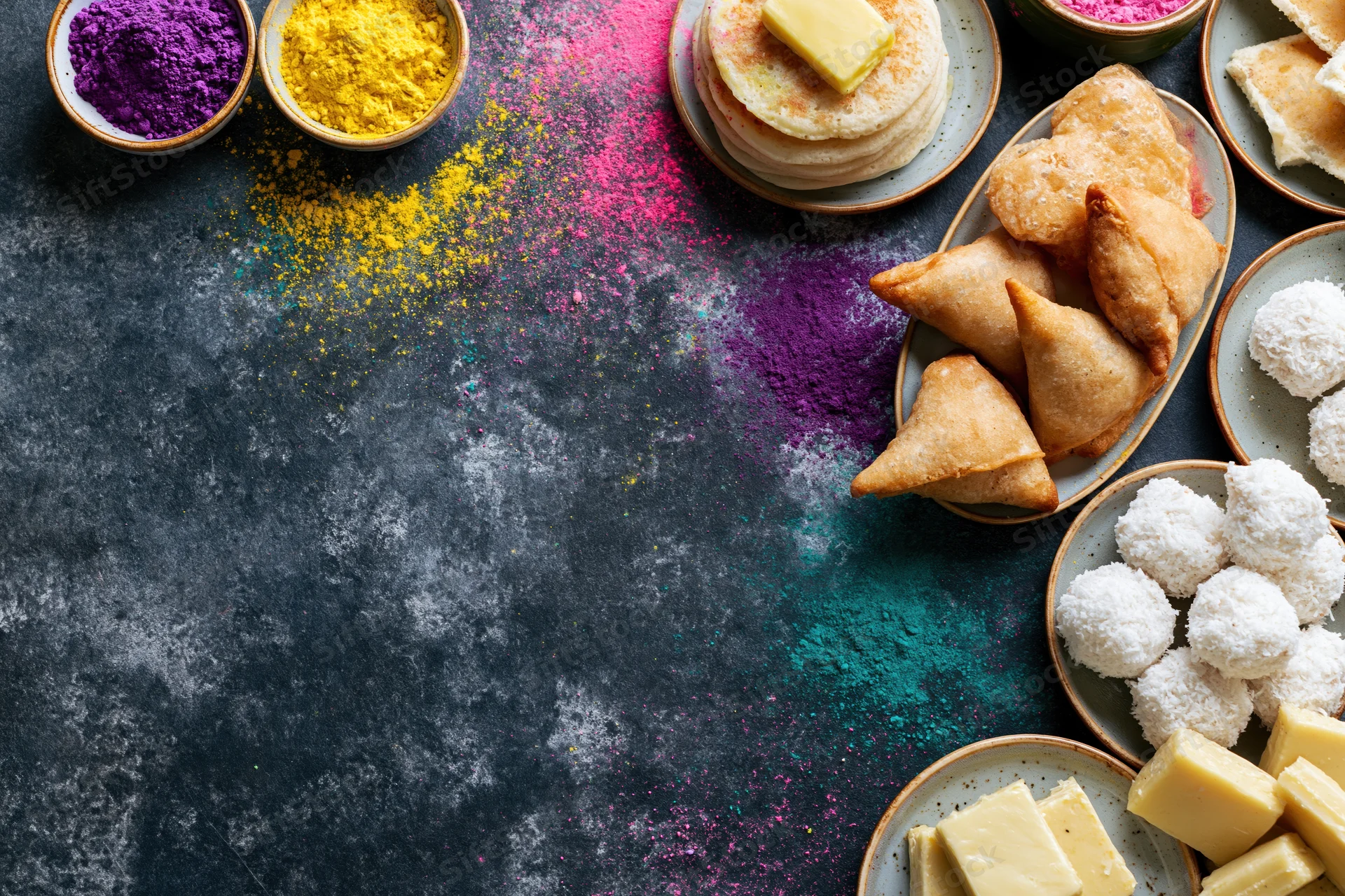 Holi festival food spread arranged along the right edge of a dark textured surface with colorful powders and traditional sweets