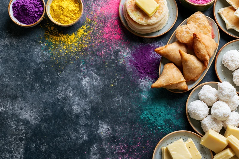 Holi festival food spread arranged along the right edge of a dark textured surface with colorful powders and traditional sweets