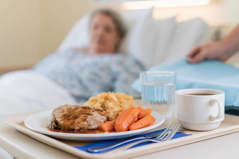 Hospital meal tray with roast meat, carrots and mashed potatoes on bedside table near patient in bed
