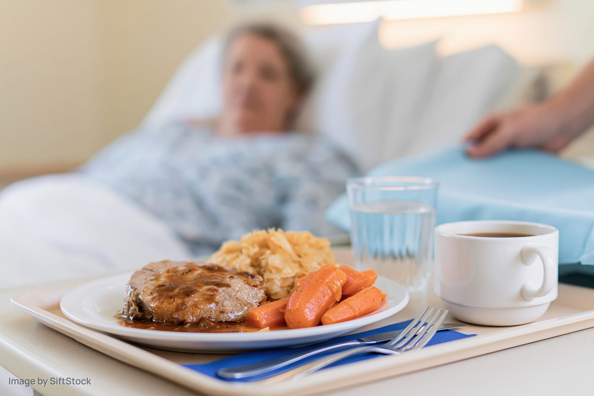 Hospital meal tray with roast meat, carrots and mashed potatoes on bedside table near patient in bed
