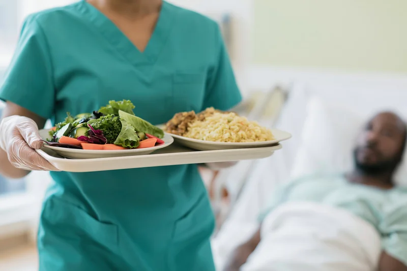 Nurse holding a meal tray with salad and rice in a hospital room near a patient in bed