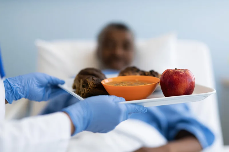 Nurse holding meal tray with soup, apple, and bread for patient in hospital bed