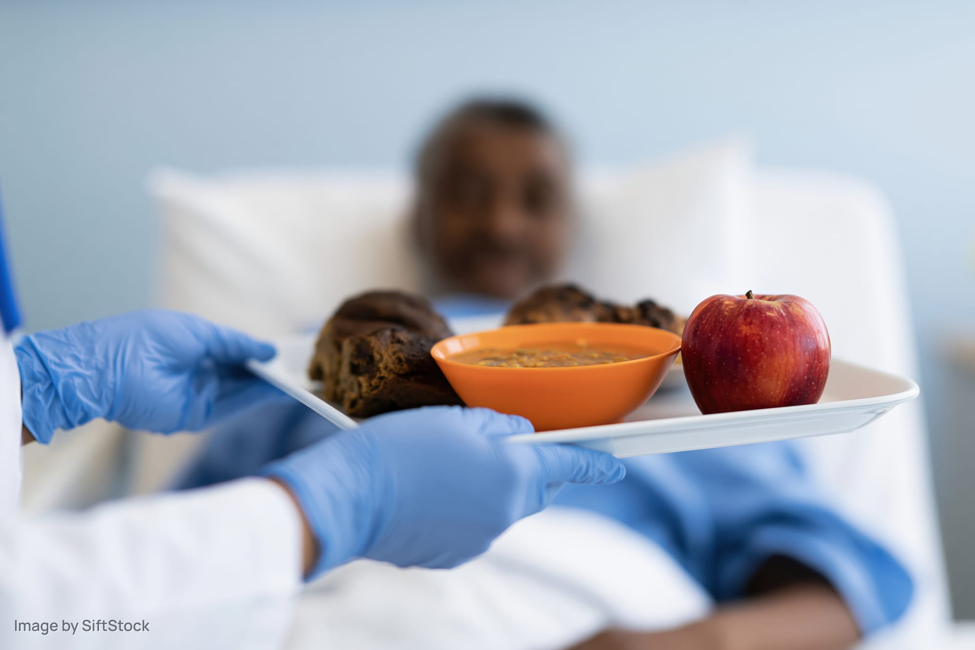 Nurse holding meal tray with soup, apple, and bread for patient in hospital bed