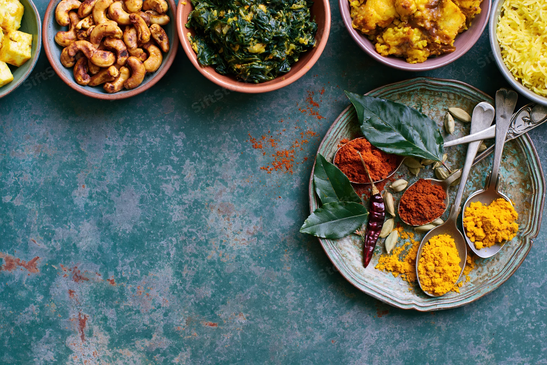 Indian food spread with spices and dishes arranged along the top edge of a dark teal surface