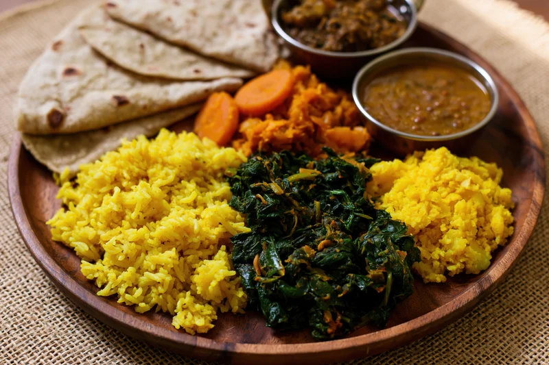 Indian thali meal served on a round wooden plate with yellow rice, spinach curry, lentil dal, roti, and vegetables on a woven cloth surface