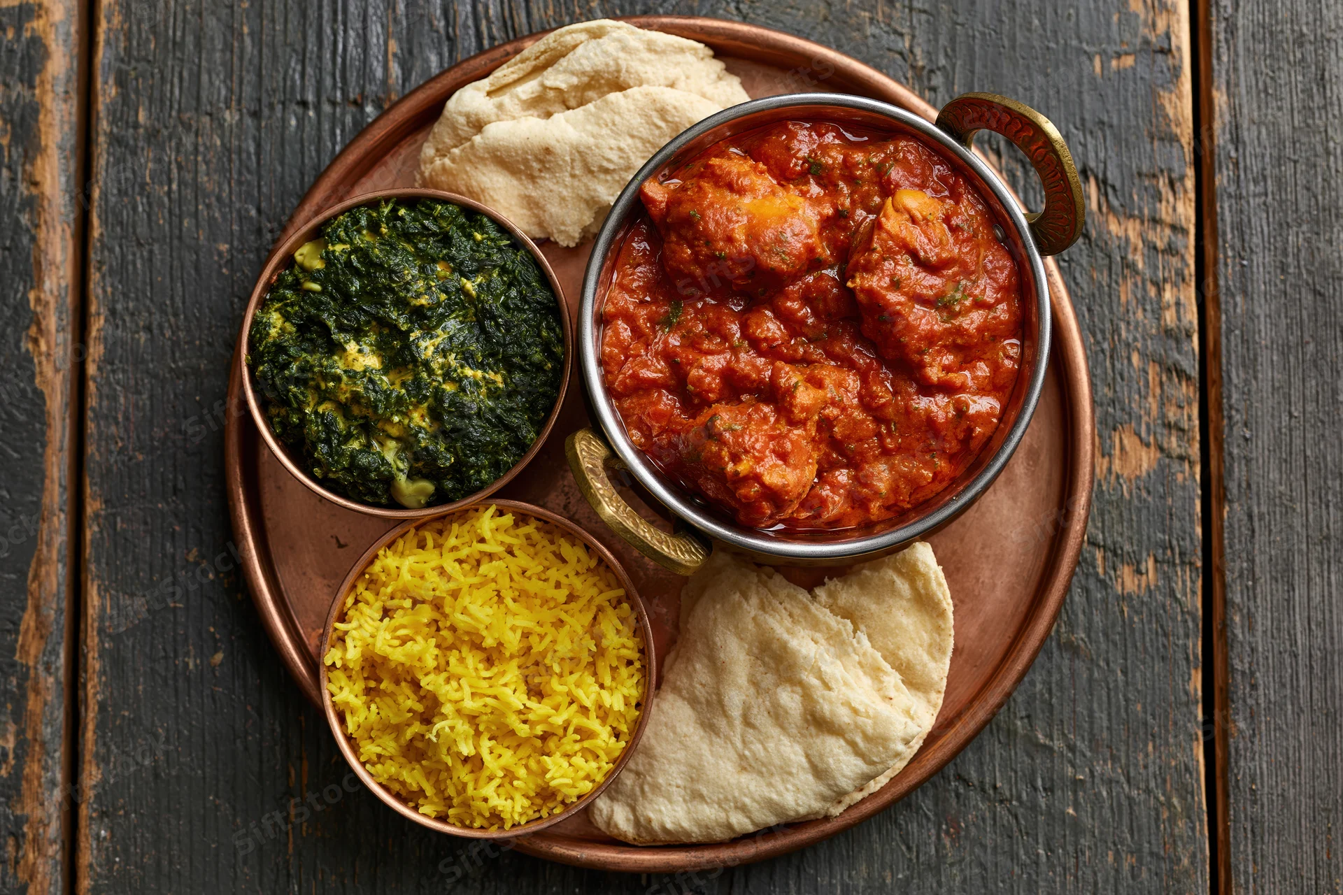 Indian thali with curry, rice, spinach, and naan on a copper tray on dark wooden table