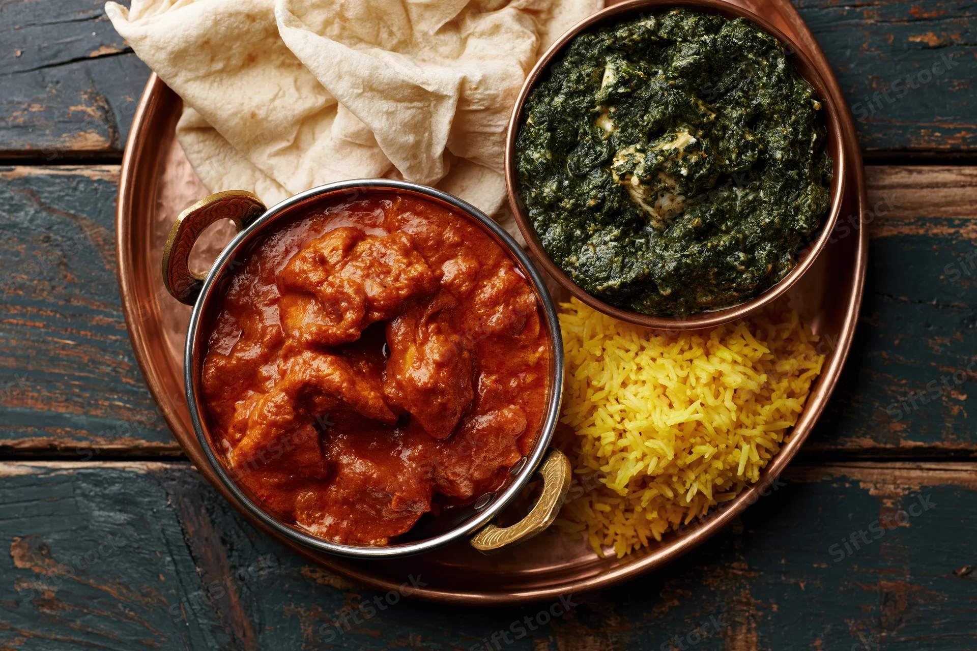 Indian thali with chicken tikka masala, palak paneer, basmati rice and naan on a copper tray on wooden table