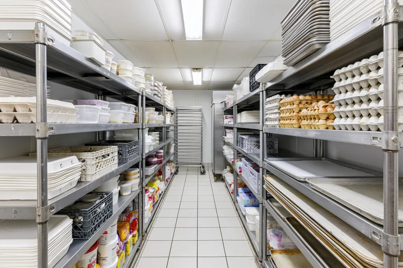 Narrow aisle between metal shelves stocked with food containers and egg cartons in a commercial kitchen walk-in refrigerator