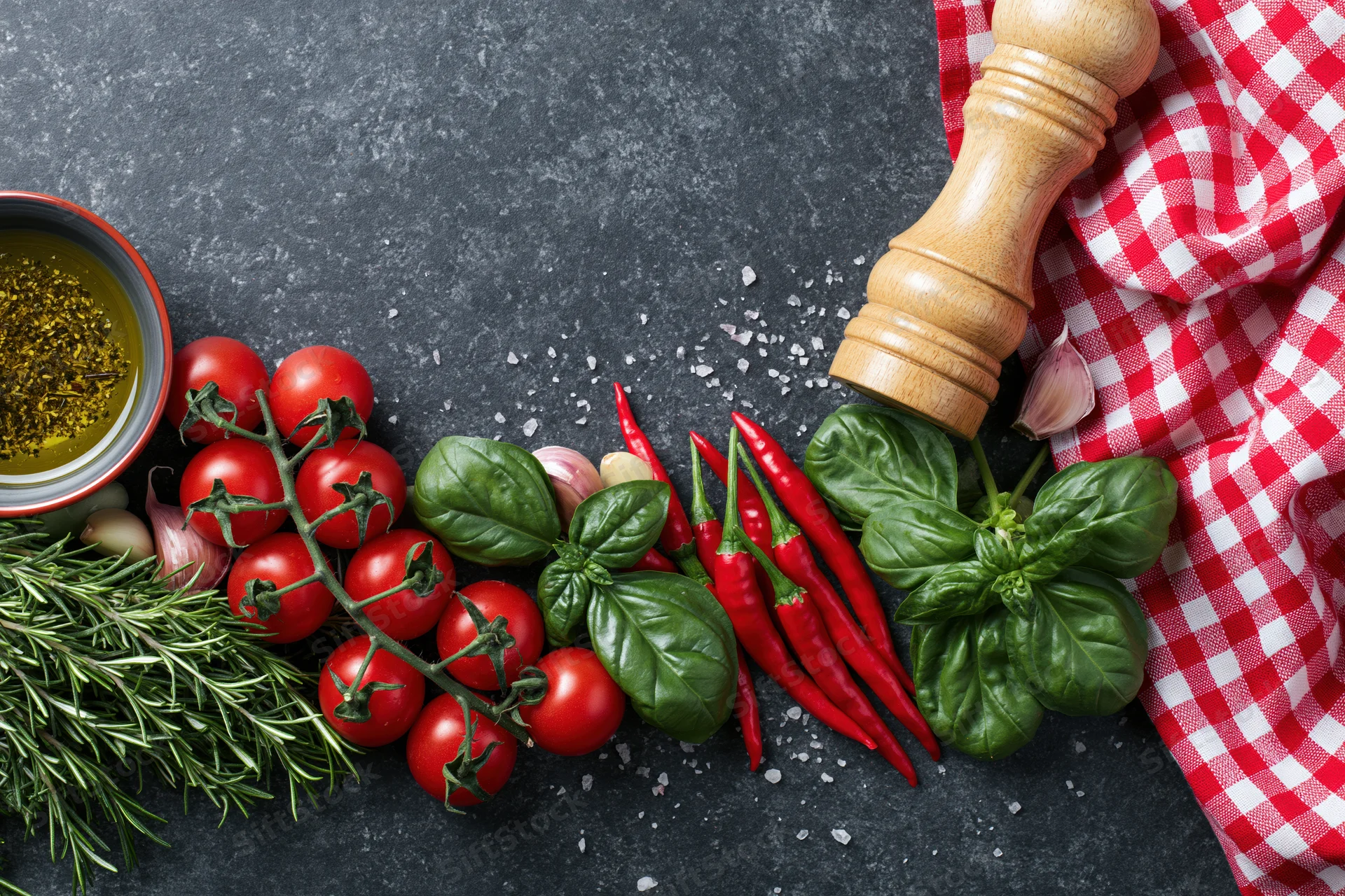 Fresh Italian cooking ingredients arranged along the bottom edge of a dark countertop with a red and white checkered cloth and wooden pepper grinder