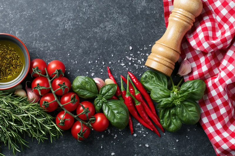 Fresh Italian cooking ingredients arranged along the bottom edge of a dark countertop with a red and white checkered cloth and wooden pepper grinder