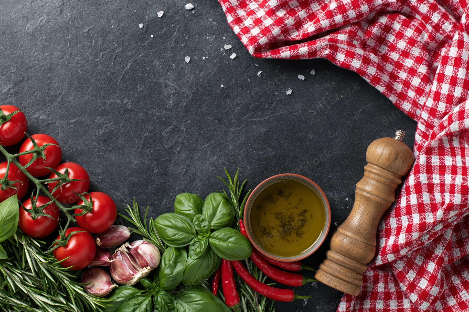 Fresh italian cooking ingredients arranged along the bottom edge of a dark slate surface with a red and white checkered cloth