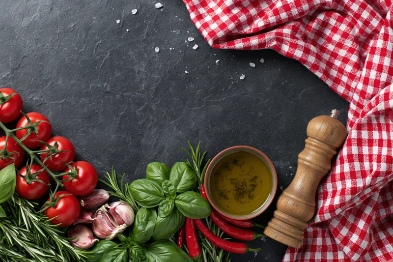 Fresh italian cooking ingredients arranged along the bottom edge of a dark slate surface with a red and white checkered cloth