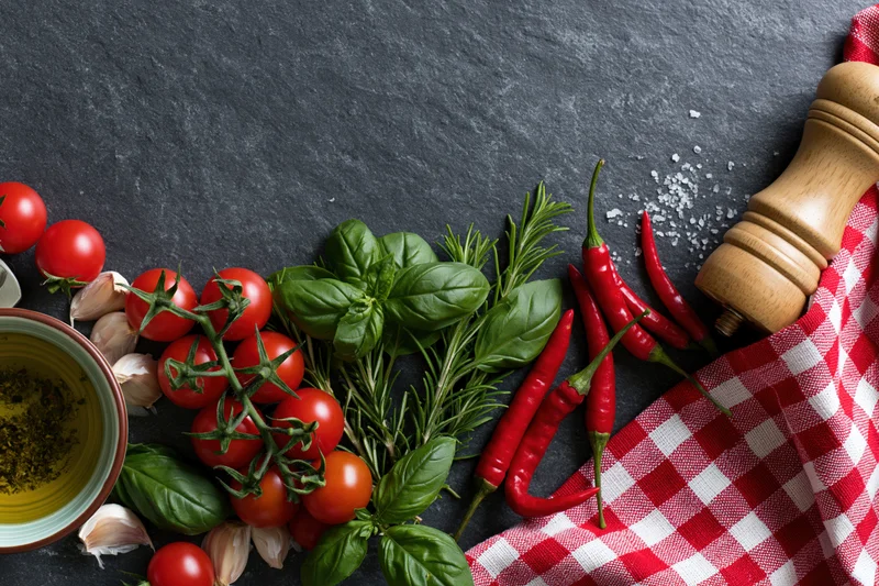 Fresh Italian cooking ingredients arranged along the bottom edge of a dark slate surface with a red checkered cloth and wooden pepper grinder
