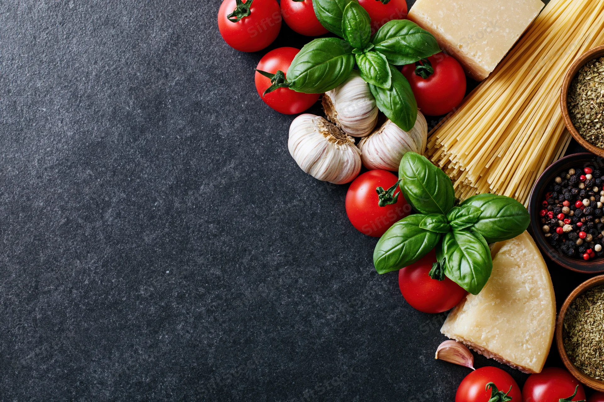 Italian cooking ingredients arranged along the right edge of a dark stone surface with copyspace on the left