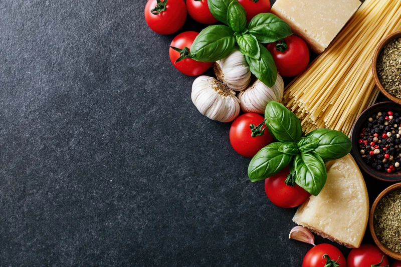 Italian cooking ingredients arranged along the right edge of a dark stone surface with copyspace on the left