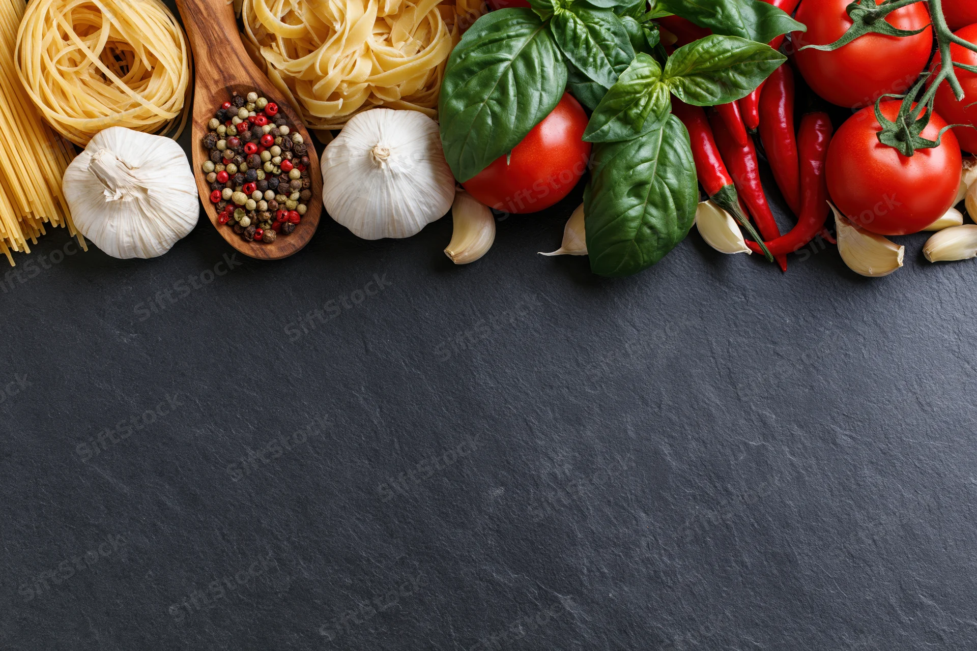 Italian cooking ingredients arranged along the top edge of a dark slate surface with copyspace below
