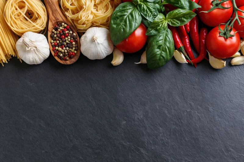 Italian cooking ingredients arranged along the top edge of a dark slate surface with copyspace below