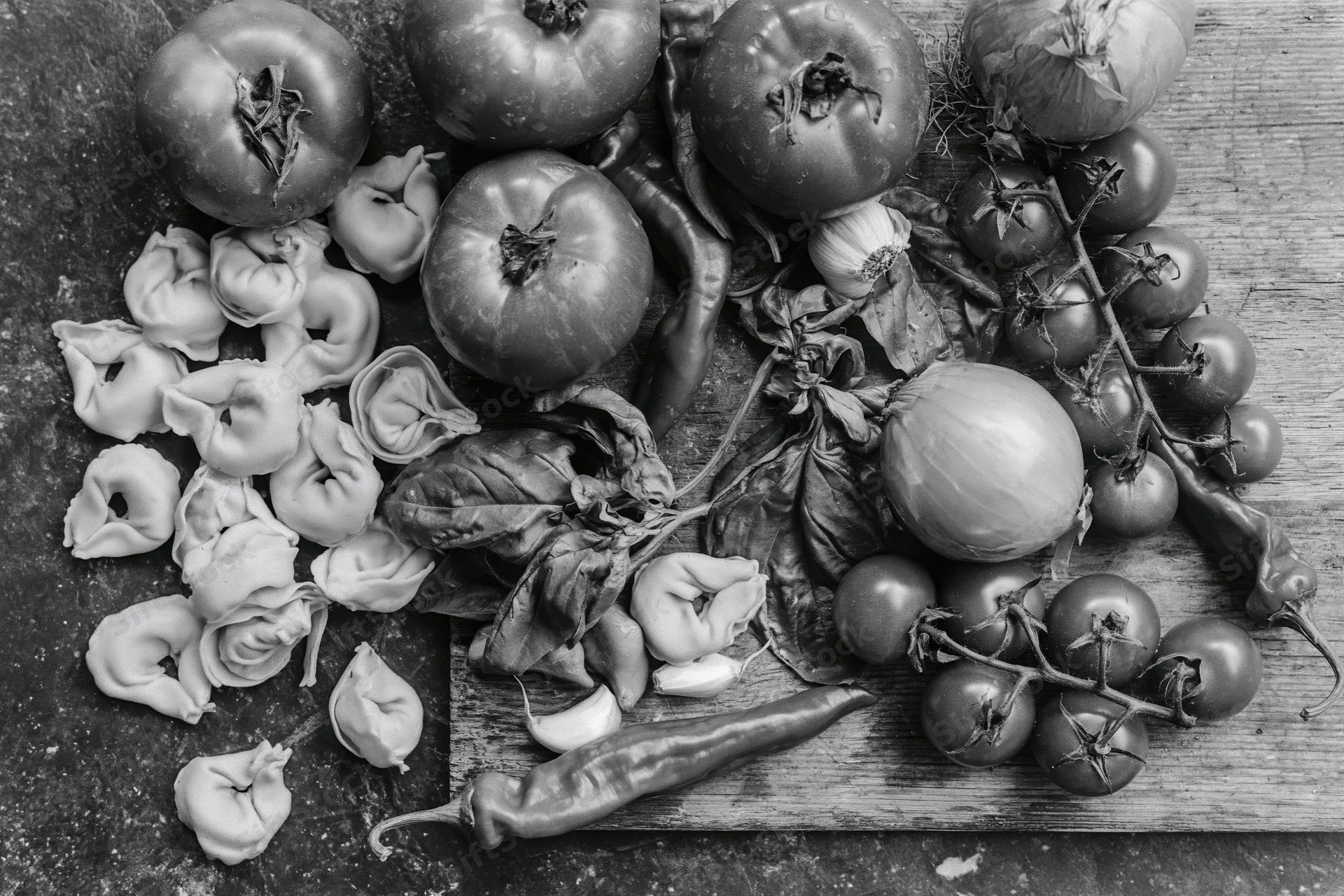 Fresh italian cooking ingredients including tomatoes onions peppers basil garlic and tortellini arranged on a dark stone surface and wooden board