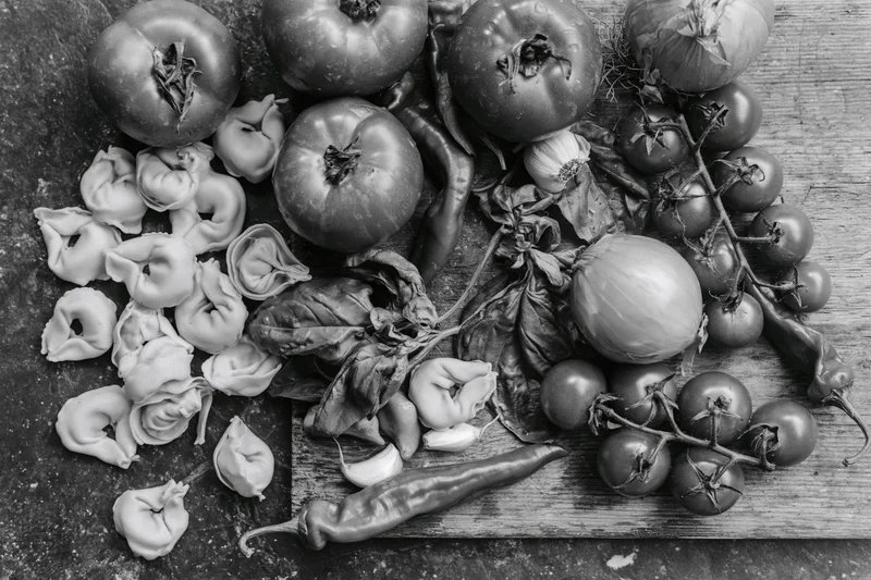 Fresh italian cooking ingredients including tomatoes onions peppers basil garlic and tortellini arranged on a dark stone surface and wooden board