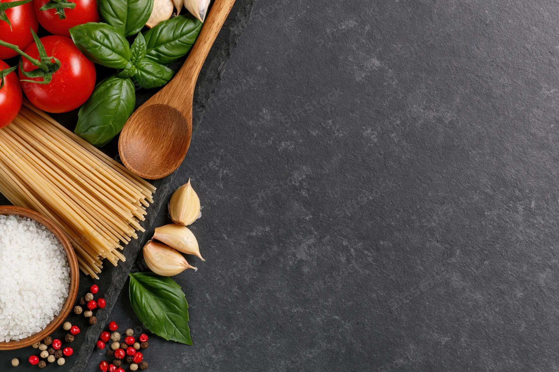 Italian pasta ingredients arranged along the left edge of a dark stone countertop with copyspace