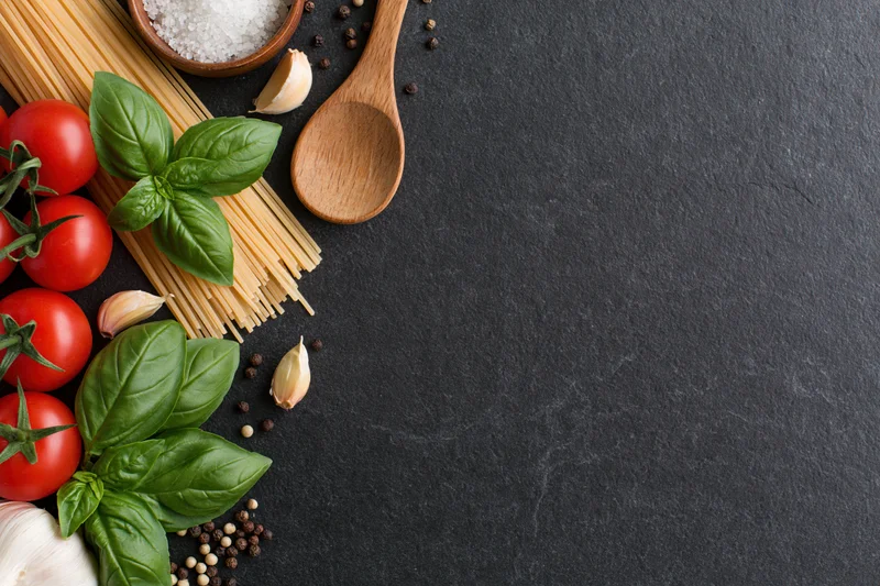 Italian pasta ingredients arranged along left edge of dark slate surface with copyspace