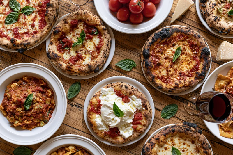 Overhead view of multiple pizzas and Italian dishes arranged on a wooden table with basil, cheese, and wine