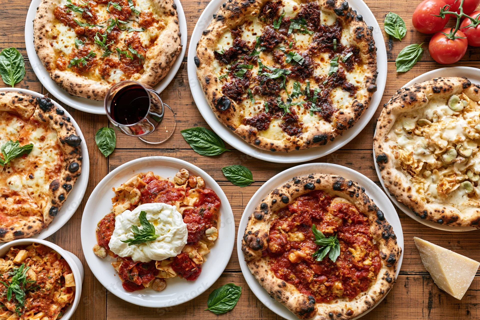 Overhead view of multiple pizzas and pasta dishes arranged on a wooden table with fresh basil, tomatoes, and wine