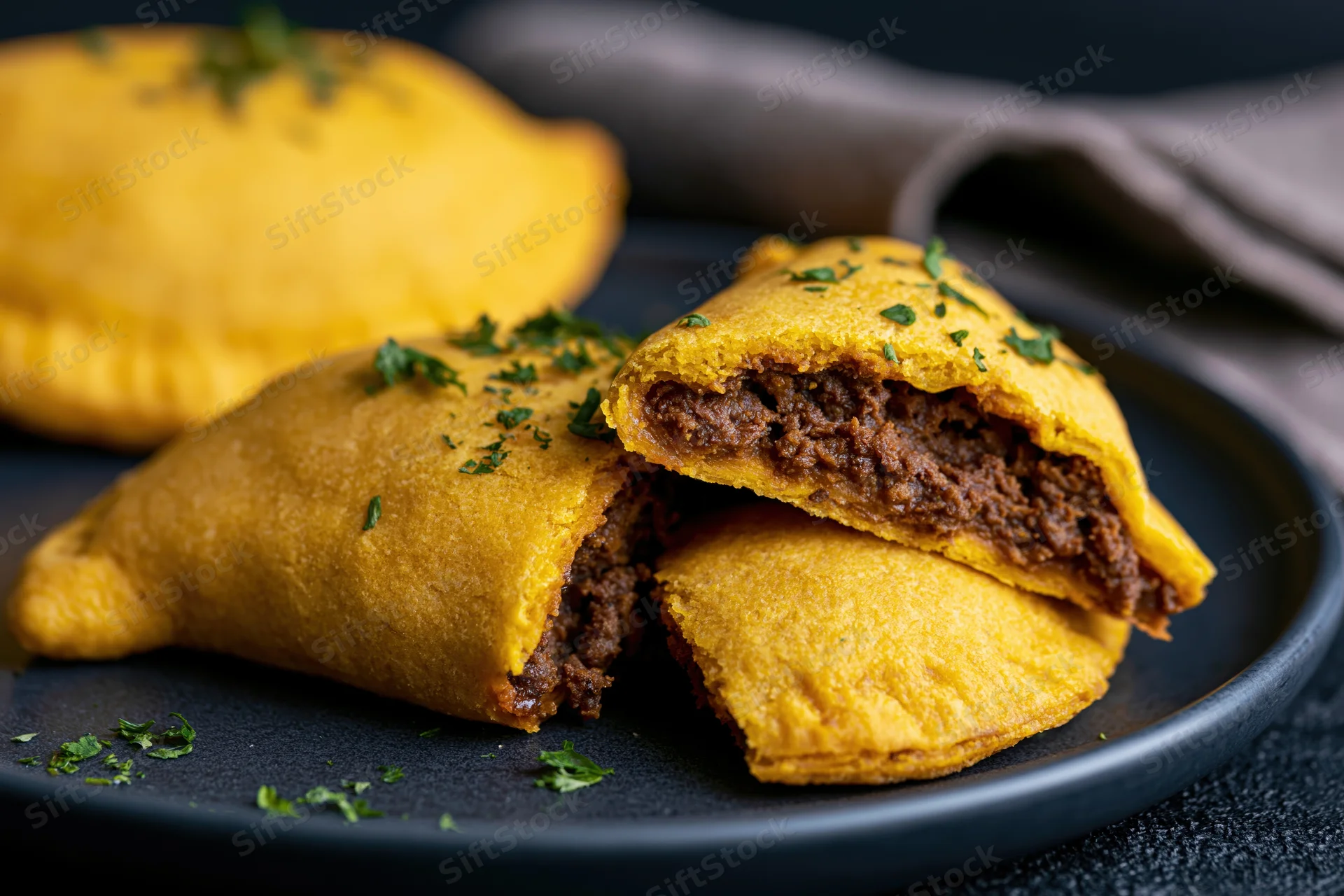 Jamaican beef patties on a dark ceramic plate with one cut open to show meat filling and garnished with parsley