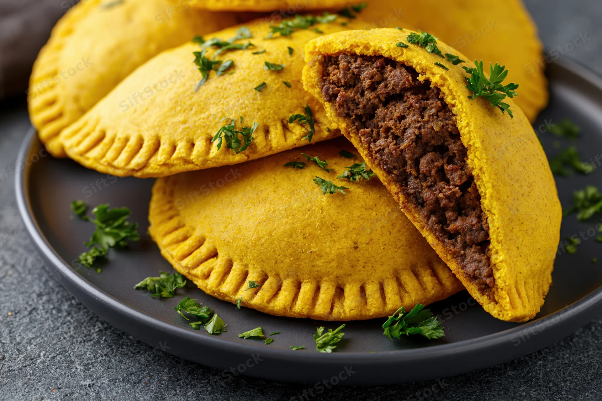 Jamaican beef patties stacked on a dark ceramic plate with one cut open to show meat filling and garnished with parsley