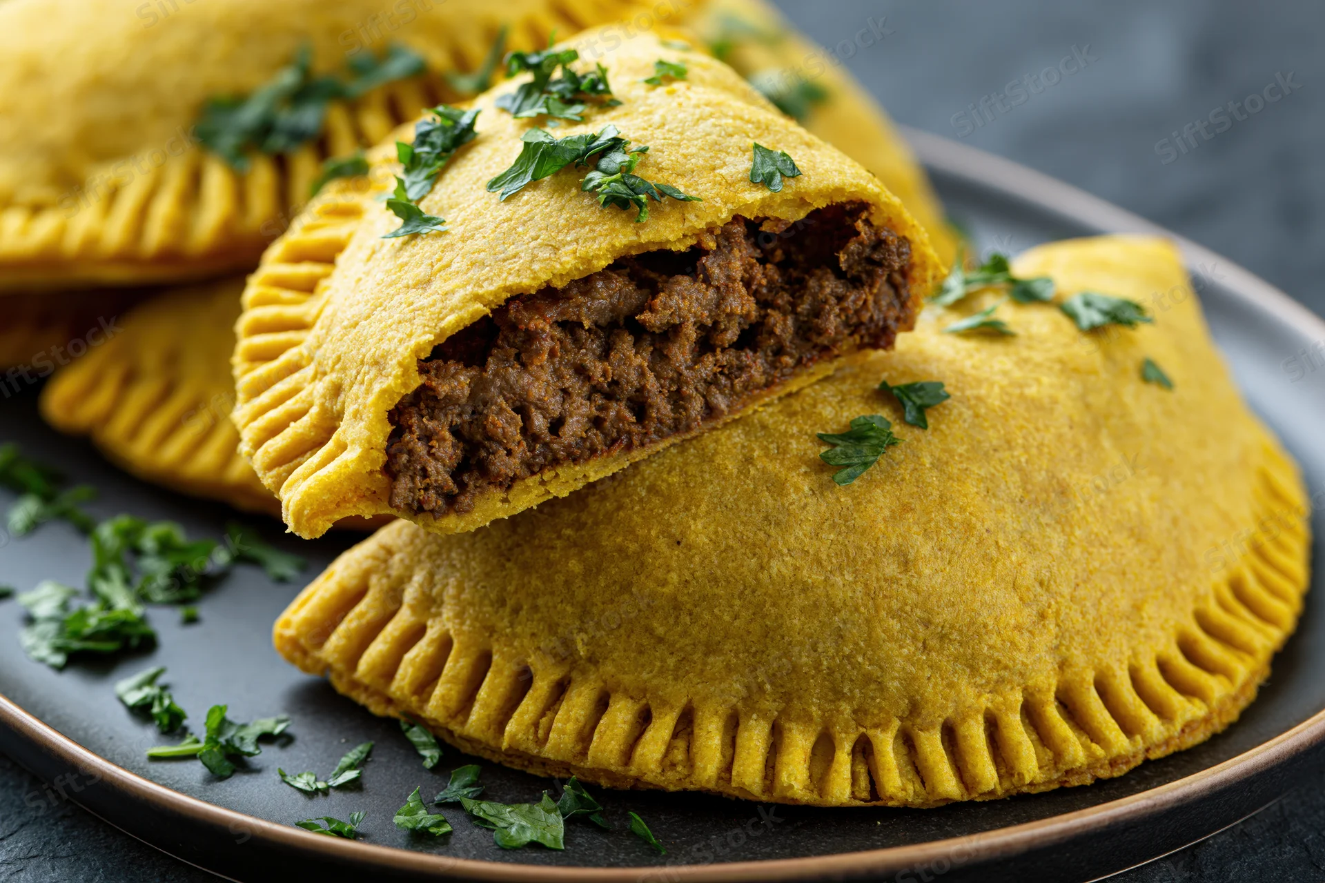 Jamaican beef patties stacked on a dark ceramic plate with one cut open to show meat filling and garnished with parsley