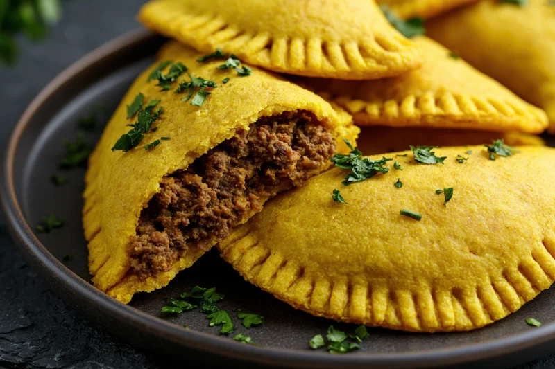 Jamaican beef patties stacked on a dark ceramic plate with one cut open to show meat filling and garnished with parsley