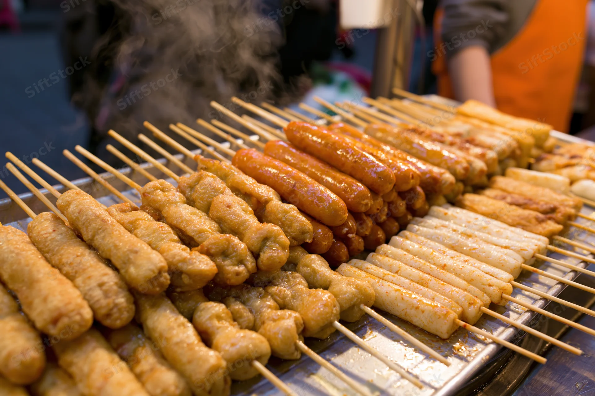 Rows of grilled korean street food skewers steaming on a metal tray at a night market stall