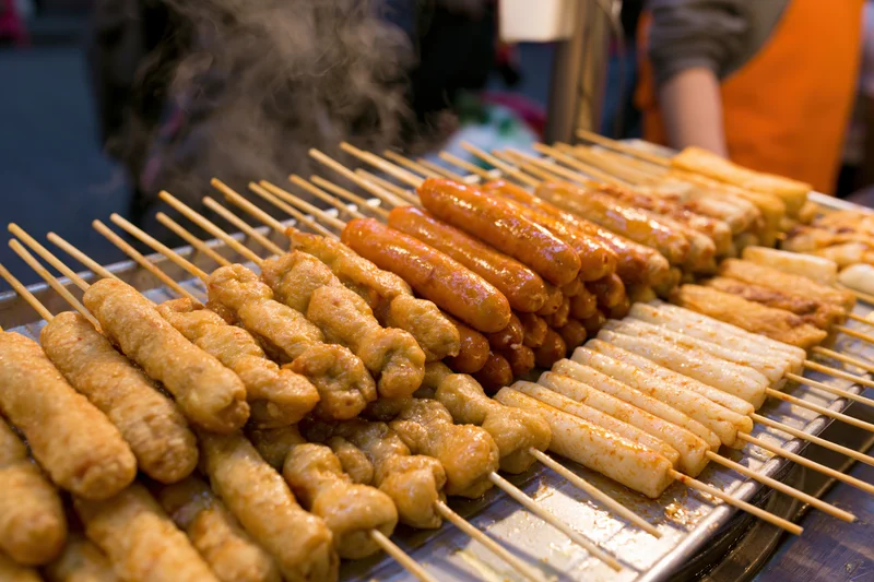 Rows of grilled korean street food skewers steaming on a metal tray at a night market stall