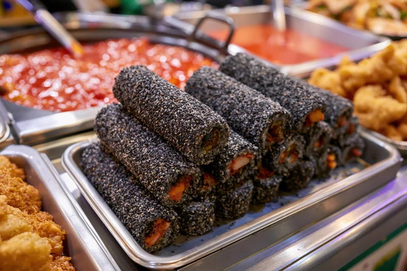 Black sesame-covered kimbap rolls stacked on a metal tray at a korean street food stall counter