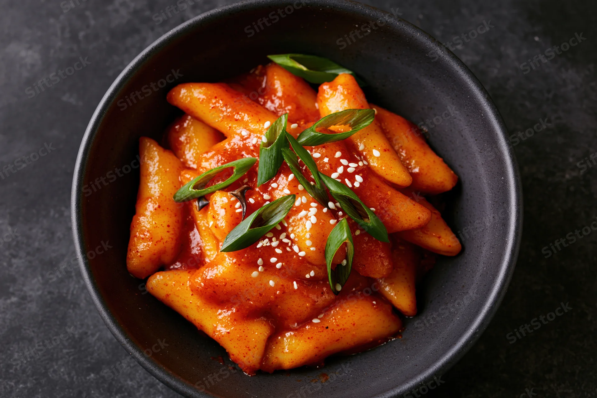 Tteokbokki rice cakes in spicy gochujang sauce served in a black bowl with sesame seeds and green onions
