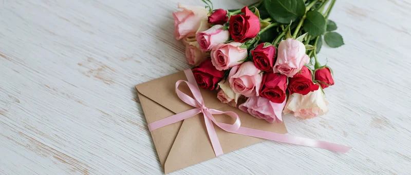 Bouquet of pink and red roses with a kraft envelope tied by a pink ribbon on a white wooden surface