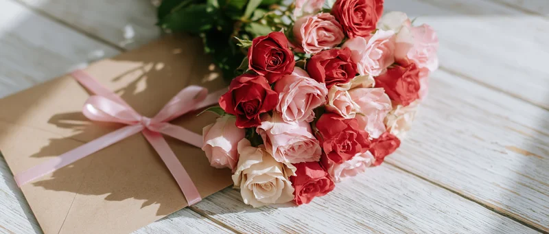 Bouquet of pink red and cream roses beside a kraft paper envelope tied with a pink satin ribbon on a wooden table