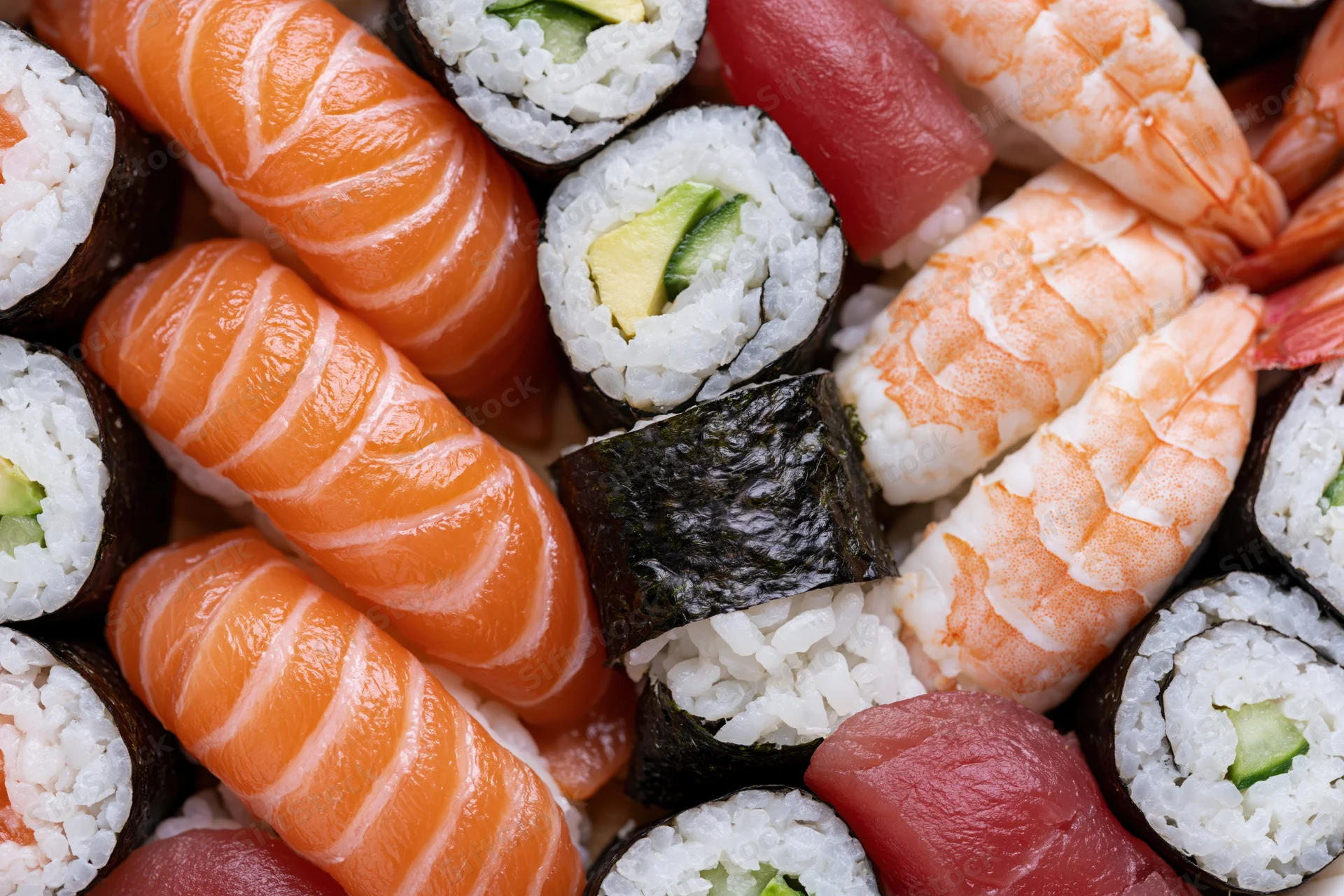 Closeup of assorted sushi pieces including salmon nigiri, shrimp tempura, tuna sashimi, and avocado maki arranged tightly together