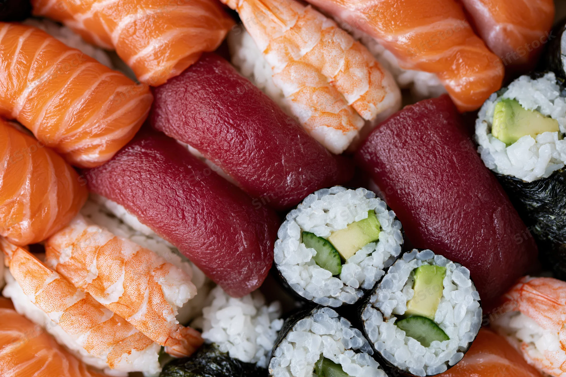 Closeup of assorted sushi pieces including salmon nigiri, tuna nigiri, shrimp nigiri, and avocado cucumber maki arranged tightly together