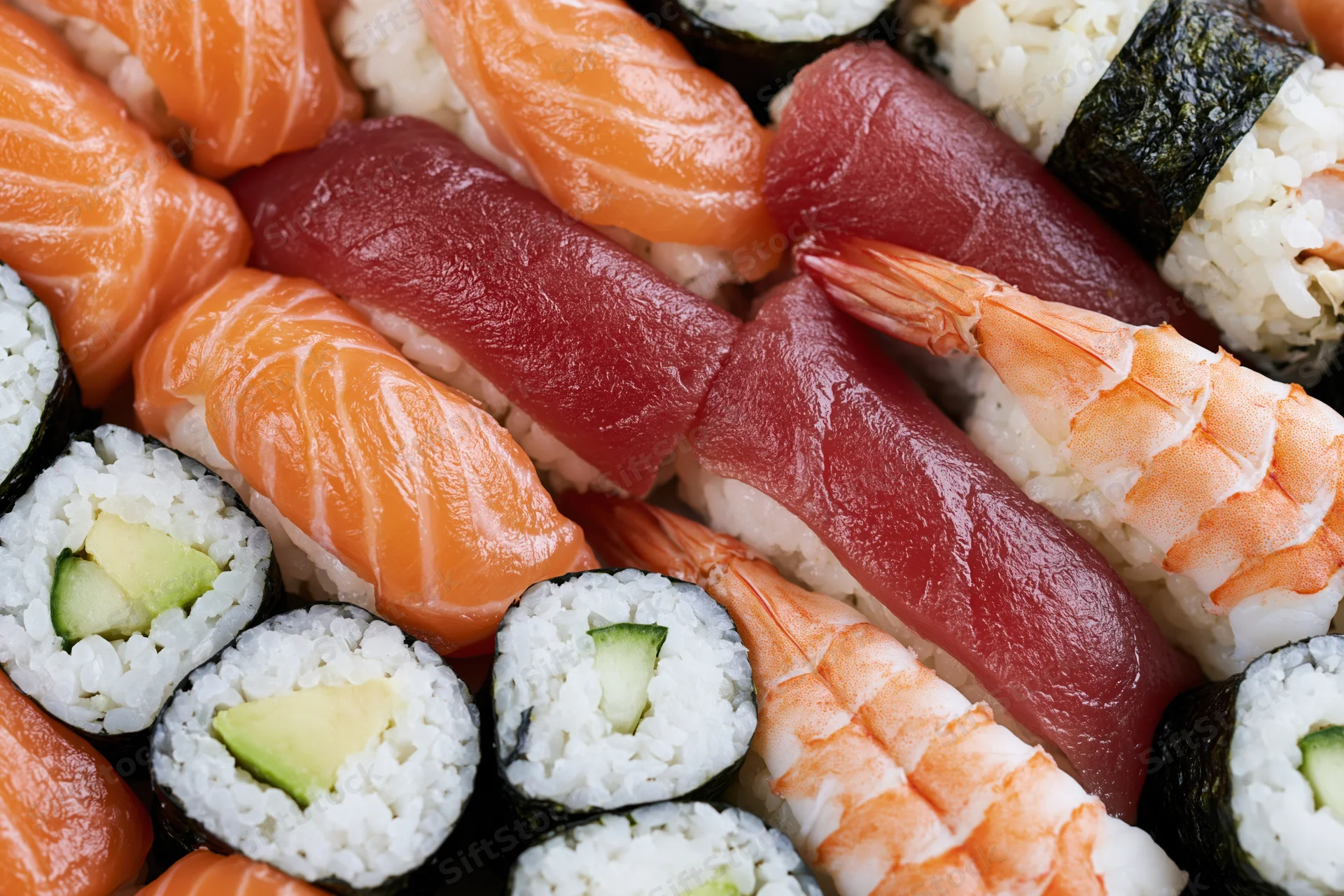 Closeup of assorted sushi pieces including salmon, tuna, shrimp, and avocado maki arranged tightly on a plate