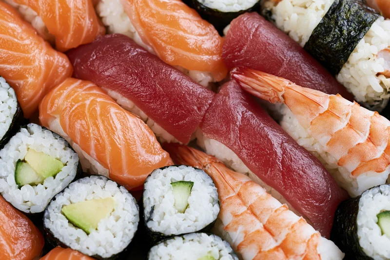 Closeup of assorted sushi pieces including salmon, tuna, shrimp, and avocado maki arranged tightly on a plate