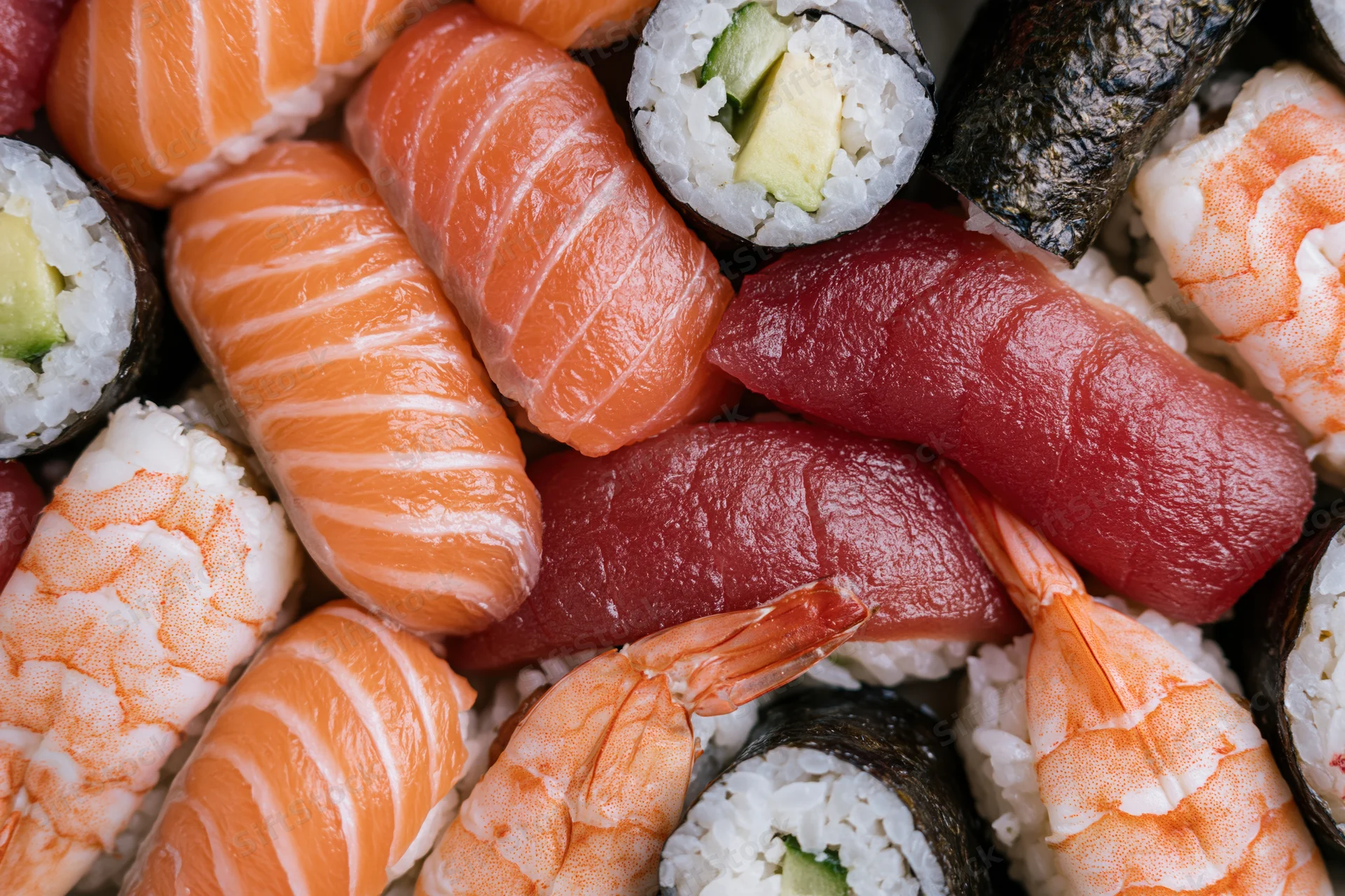Closeup of assorted sushi pieces including salmon, tuna, shrimp, and avocado maki arranged tightly together