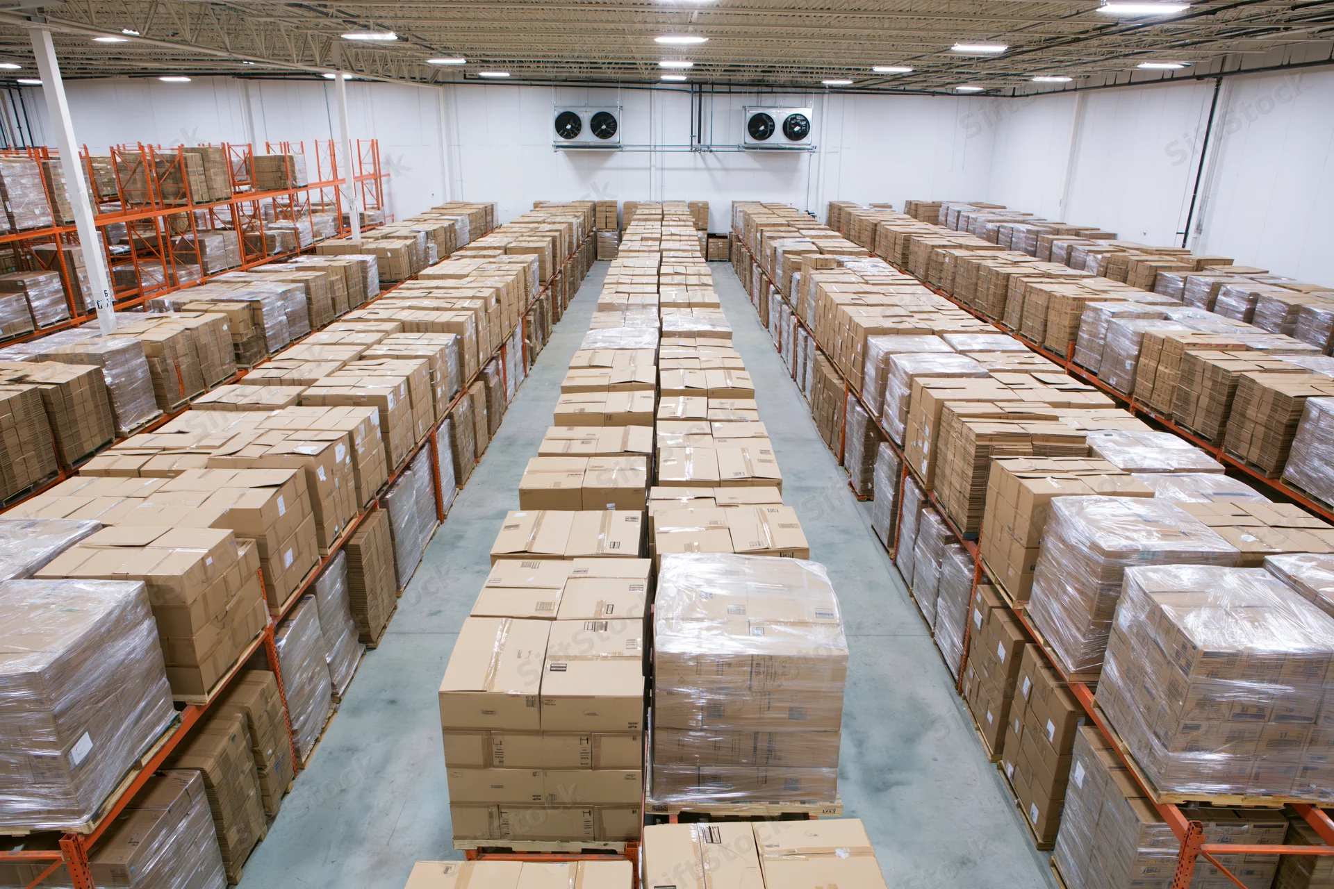Rows of stacked cardboard boxes on pallets and shelves in a large warehouse interior