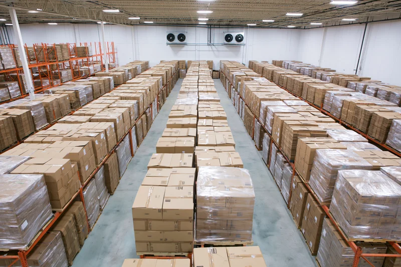 Rows of stacked cardboard boxes on pallets and shelves in a large warehouse interior