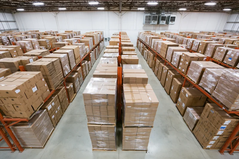 Rows of stacked cardboard boxes on pallets and metal shelves inside a large warehouse interior