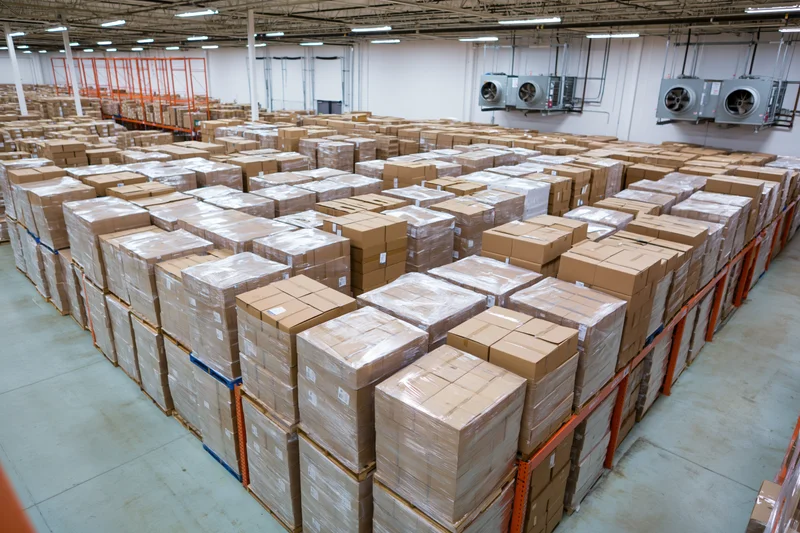 Rows of stacked cardboard boxes on pallets in a large commercial warehouse interior