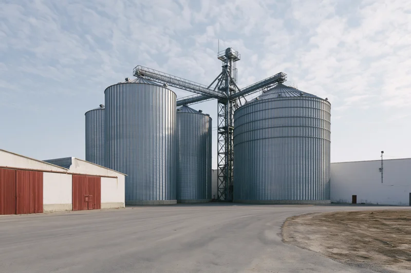 Large grain elevator complex with multiple tall corrugated metal silos and conveyor system under a partly cloudy sky
